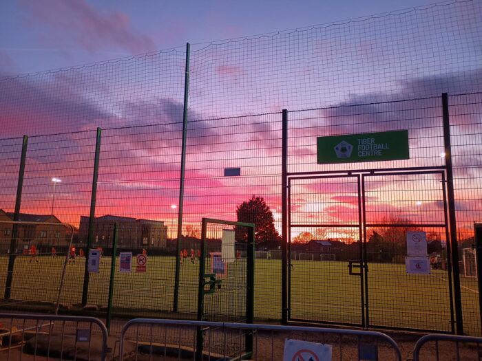 Sunset sky behind a football pitch fence.